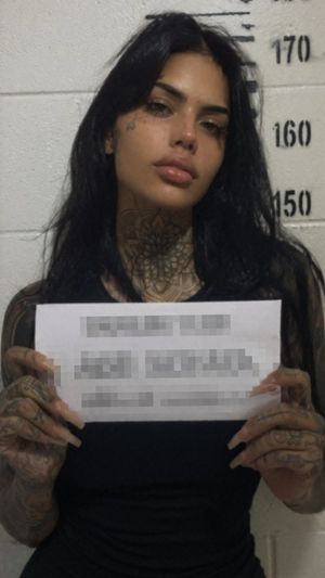 Generates a realistic, gritty mugshot of a heavily tattooed woman holdin...